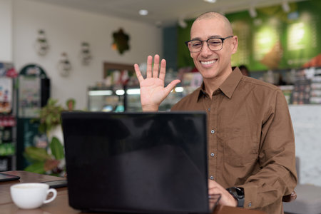 Mature Hispanic man using laptop in indoor cafe, wearing eyeglasses and button down shirt. Confident and focused expression. Remote work, freelance, tech-savvy lifestyle concept in urban setting.の写真素材