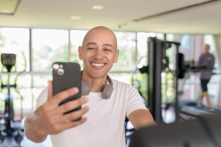 Bald Hispanic man working out on a stationary exercise bike inside modern gym while using phone. Focused fitness and workout concept showing strength, cardio, and motivation.の写真素材