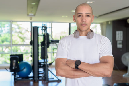 Bald Latino man standing inside modern gym with confident expression. Portraying wellness, training discipline, and a strong healthy lifestyle in indoor environment.の写真素材