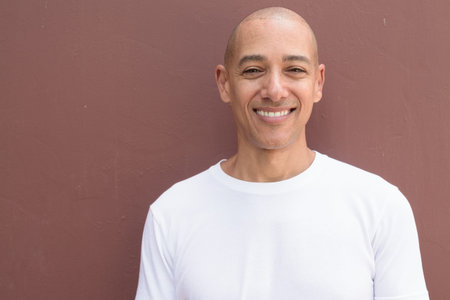 Bald Latin man against wall in casual white t-shirt. Minimalist, style emphasizing confidence,...