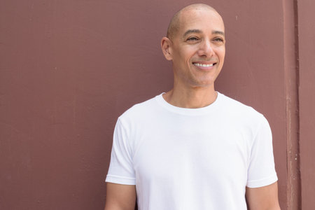 Bald Latin man against wall in casual white t-shirt. Minimalist, style emphasizing confidence, identity and wellness lifestyle.の写真素材