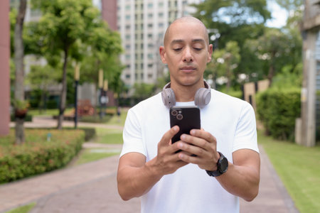 Bald Hispanic man using phone while walking in park or urban area. Represents modern digital lifestyle, healthy movement habits, and confident outdoor balance.の写真素材