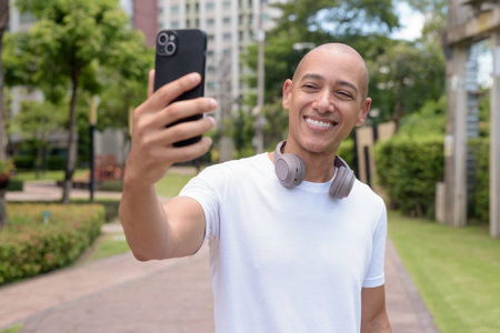 Bald Hispanic man using phone while walking in park or urban area. Represents modern digital lifestyle, healthy movement habits, and confident outdoor balance.の写真素材