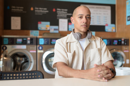 Bald Hispanic man in casual outfit sitting in self-service laundry shop. Urban portrait with washing machines behind him, showing everyday city lifestyle.の写真素材