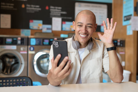 Bald Hispanic man in casual clothes sitting in self-service laundry shop using smartphone. Everyday...