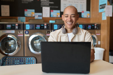 Bald Hispanic man in casual clothes sitting in laundry shop using laptop computer. Everyday urban lifestyle concept with washing machines in background.の写真素材