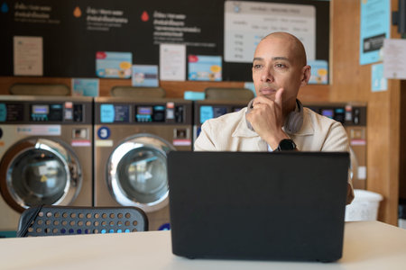 Bald Hispanic man in casual clothes sitting in laundry shop using laptop computer. Everyday urban...