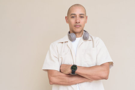 Portrait of bald Hispanic man wearing casual beige shirt and headphones around neck, standing...