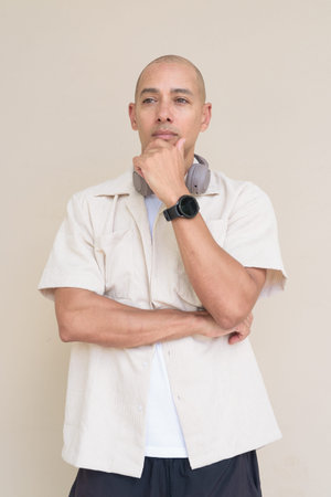 Portrait of bald Hispanic man wearing casual beige shirt and headphones around neck, standing...
