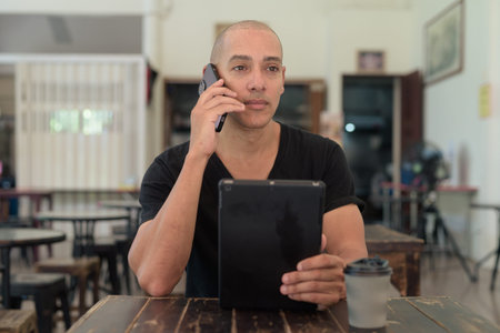 Portrait of bald Latin man wearing black t-shirt, sitting in simple outdoors coffee shop restaurant aloneの写真素材