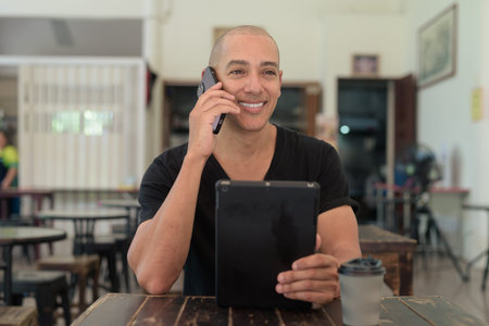 Portrait of bald Latin man wearing black t-shirt, sitting in simple outdoors coffee shop restaurant...