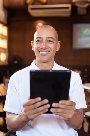 Portrait of bald Latin man wearing white polo shirt, sitting in stylish Chinese style coffee shop,...