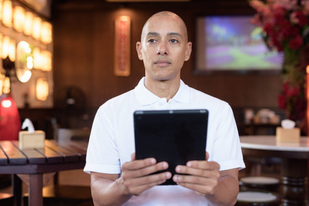Portrait of bald Latin man wearing white polo shirt, sitting in stylish coffee shop, looking...