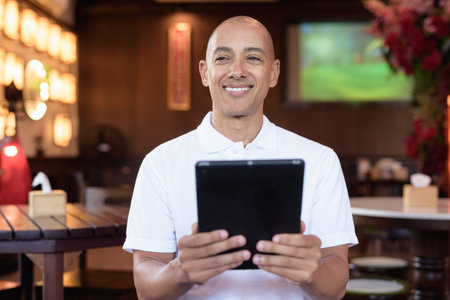 Portrait of bald Latin man wearing white polo shirt, sitting in stylish Chinese style coffee shop, looking confident in natural light indoor setting.の写真素材