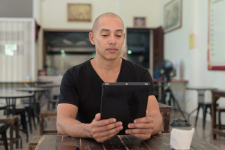 Bald Hispanic man sitting in outdoors cafe restaurant using digital tablet computer with focused expressionの写真素材