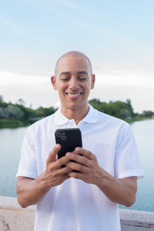 Stylish bald man wearing a white polo shirt standing by a calm lake using smartphone. Confident and relaxed expression in a natural light outdoor portrait with peaceful waterfront scenery.の写真素材