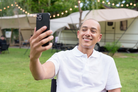 Stylish bald man in a white polo shirt relaxing at a campsite beside a camper van. Outdoor travel and camping lifestyle concept with grassy surroundings and natural light.の写真素材