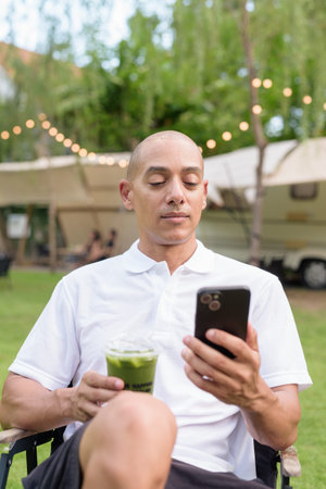 Stylish bald man in a white polo shirt relaxing at a campsite beside a camper van. Outdoor travel...