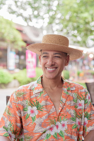 Portrait of a middle-aged Hispanic man in a Panama hat and tropical short-sleeve shirt sitting at an outdoor cafe table on a sunny day. The male tourist looks relaxed and confident.の写真素材