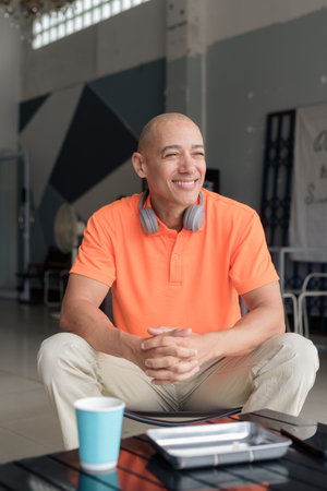 Handsome middle aged bald man in orange polo shirt sitting at cafe table with coffee cup and wearing headphones happily smiling. Casual lifestyle concept.の写真素材