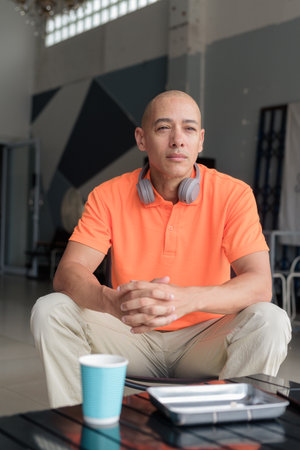 Middle aged man in orange polo shirt sitting at cafe table with coffee cup and wearing headphones while thinking. Casual lifestyle concept.の写真素材
