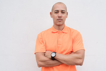 Bald Italian man in orange polo shirt studio portrait against white wall with arms crossed