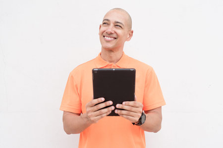 Studio portrait of handsome middle aged bald Hispanic man in orange polo shirt against plain white...