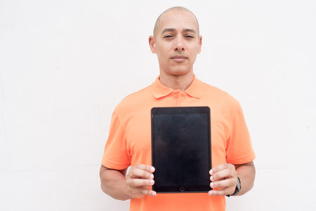 Studio portrait of handsome middle aged bald Hispanic man in orange polo shirt against plain white wall background using digital tablet computer. Casual lifestyle and fashion concept.の写真素材