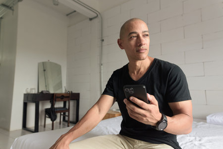 Middle aged Hispanic bald man wearing black t-shirt sitting on bed while using phone inside minimalistic white industrial style apartment. Casual lifestyle and communication at home.の写真素材