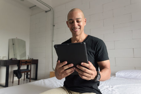 Bald man wearing black t-shirt sitting on bed while using digital tablet computer inside...