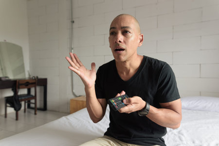 Middle aged man in black t-shirt sitting on bed while watching television in a minimalistic white...