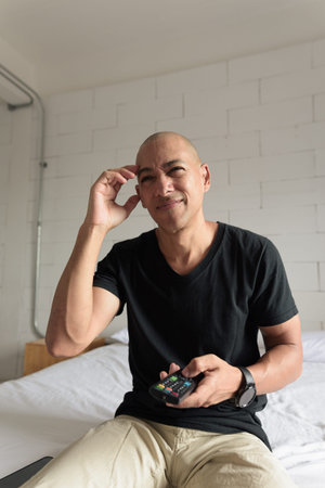 Middle aged man in black t-shirt sitting on bed while watching television in a minimalistic white...