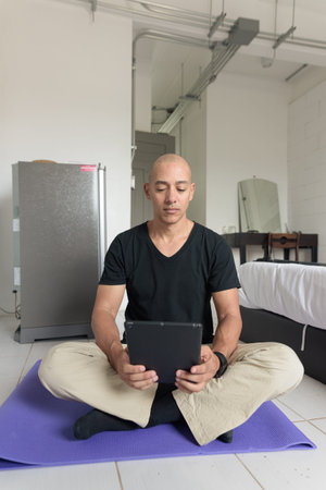 Middle aged bald man in black t-shirt sitting cross-legged on yoga mat while doing yoga and...