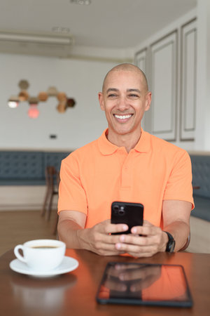 Handsome middle aged bald man in orange polo shirt at cafe table with coffee cup and using...