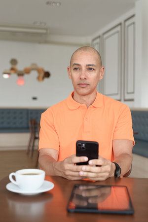 Handsome middle aged bald man in orange polo shirt at cafe table with coffee cup and using...