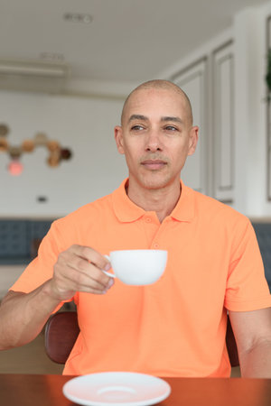 Handsome middle aged bald man in orange polo shirt sitting at cafe table holding coffee cup. Casual...