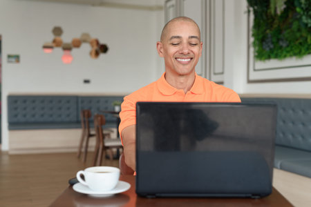 Handsome middle aged bald man in orange polo shirt at cafe table with coffee cup working on laptop...