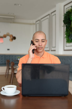 Handsome middle aged bald man in orange polo shirt at cafe table with coffee cup working on laptop...