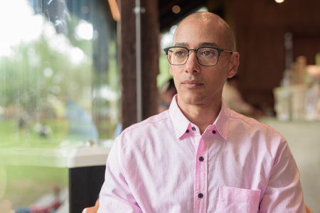 Bald Hispanic mature male in 40s wearing pink casual business shirt and eyeglasses inside modern coffee shop restaurant, Concept of urban lifestyle, leisure, cafe culture, and social relaxation.の写真素材