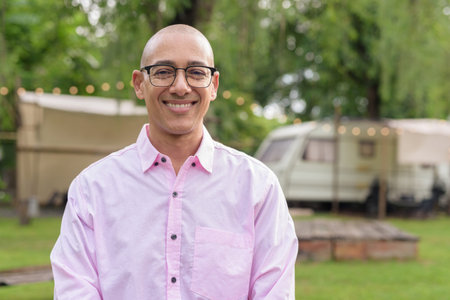 Bald Hispanic mature male in 40s wearing pink shirt and eyeglasses outdoors with camping park environment and caravan background. Concept of travel, tourism, and lifestyle in outdoor leisure setting.の写真素材