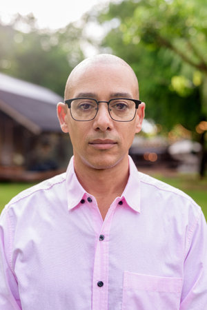 Bald Hispanic mature male in 40s wearing pink shirt and eyeglasses outdoors with camping park environment and caravan background. Concept of travel, tourism, and lifestyle in outdoor leisure setting.の写真素材