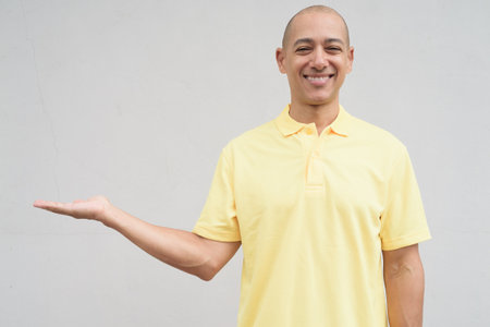 Studio portrait of a handsome mature happy man with shaved head smiling wearing yellow polo shirt, standing in front of a neutral plain white wall with copy space.の写真素材