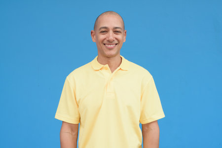 Studio portrait of a mature man with shaved head wearing yellow polo shirt, standing in front of a neutral plain blue wall with copy space. Clean isolated cut out look.の写真素材