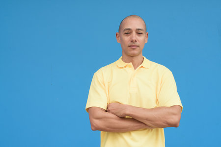 Studio portrait of a handsome mature Italian man with shaved head wearing yellow polo shirt, standing in front of a neutral plain blue wall with copy space. Clean isolated cut out look.の写真素材