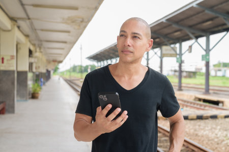 Portrait of a mature bald Hispanic man tourist in a black t-shirt using phone at a train station platform, waiting for a train while traveling in Thailand. Tourism, commuting, and transport theme.の写真素材