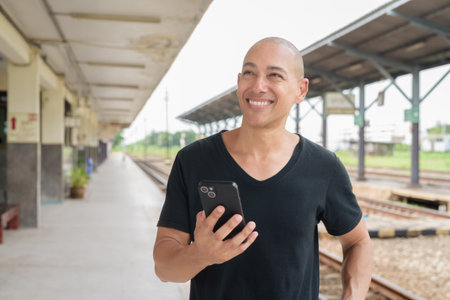 Portrait of a mature bald Hispanic man tourist in a black t-shirt using phone at a train station platform, waiting for a train while traveling in Thailand. Tourism, commuting, and transport theme.の写真素材