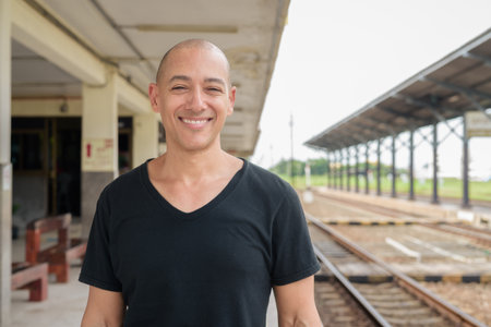 Portrait of a mature bald Hispanic man tourist in a black t-shirt standing at a train station platform, waiting for a train while traveling in Thailand.の写真素材