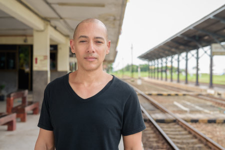 Portrait of a mature bald Hispanic man tourist in a black t-shirt standing at a train station platform, waiting for a train while traveling in Thailand.の写真素材