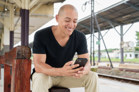 Portrait of a mature bald Hispanic man tourist in a black t-shirt sitting at a train station...