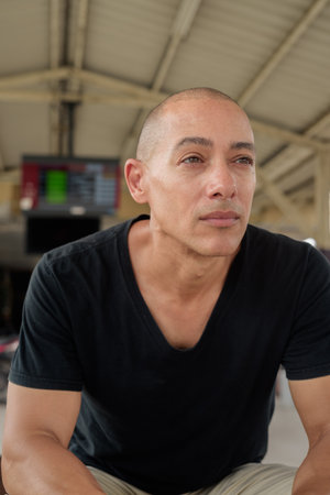Portrait of a mature bald Hispanic man tourist in a black t-shirt sitting at a train station platform, waiting for a train while traveling in Thailand. Tourism, commuting, and transport theme.の写真素材